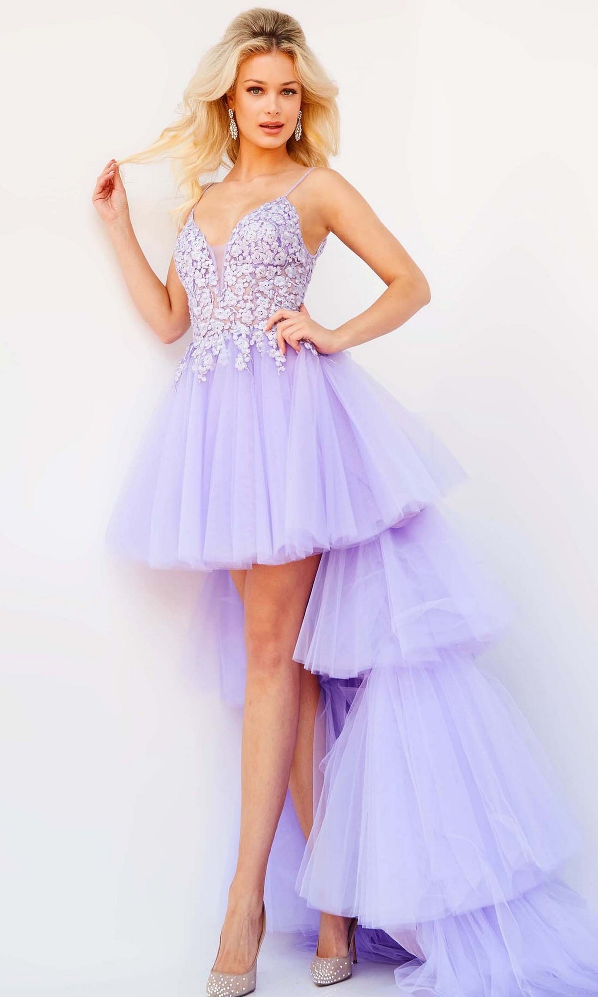 A woman poses in the Jovani Ruffled High-Low Prom Dress 07263, a light purple gown with an embroidered floral lace bodice, paired with silver high heels against a plain light background.